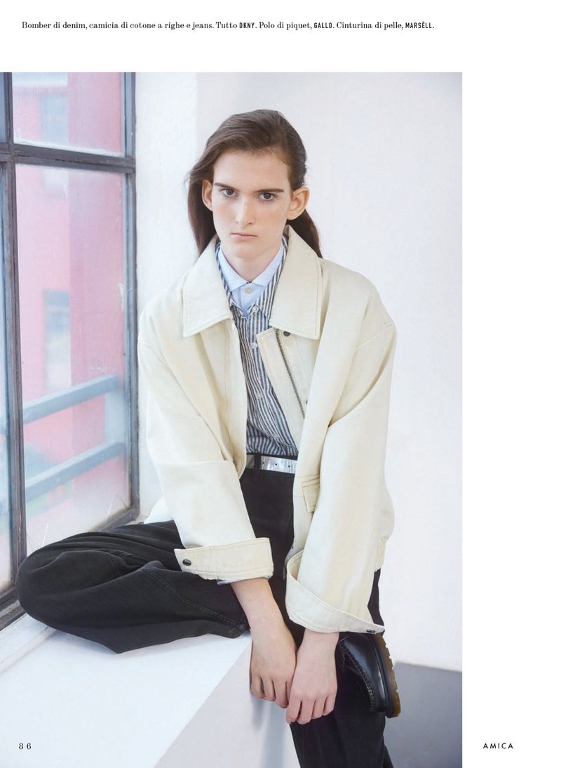 Fashion portrait of a young adult person seated by a window, wearing a light jacket, striped shirt, dark trousers and leather shoes.