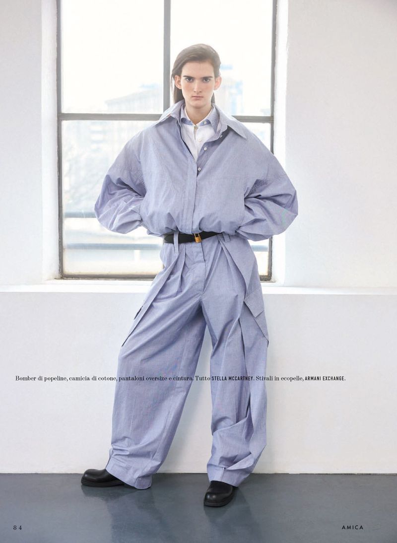 Model standing against a large window wearing a blue oversized poplin shirt, matching trousers, and dark shoes.