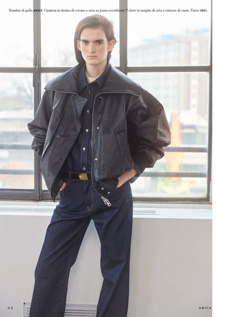 Fashionable person poses in dark denim shirt, wide-leg jeans, and a black leather bomber jacket by a window in natural light.