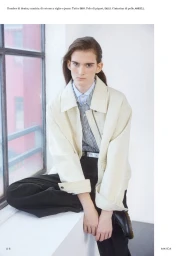 Model Tia Edney seated near a window wearing a white jacket over a striped shirt and dark trousers for Amica.