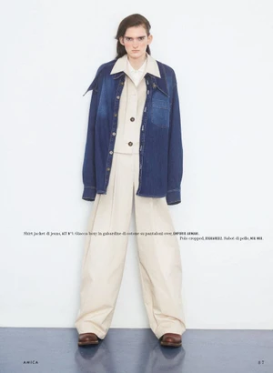 Full-length studio portrait of a model wearing an open dark denim shirt jacket over a cream button-up shirt and wide trousers.