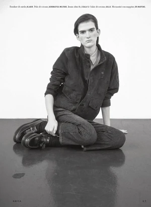 Model seated on a dark reflective studio floor against a plain background wearing a dark jacket and trousers