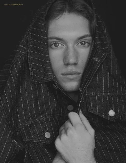 Monochrome portrait of a person with light skin wearing a striped denim jacket, styled with the jacket hood over their head, dark background.