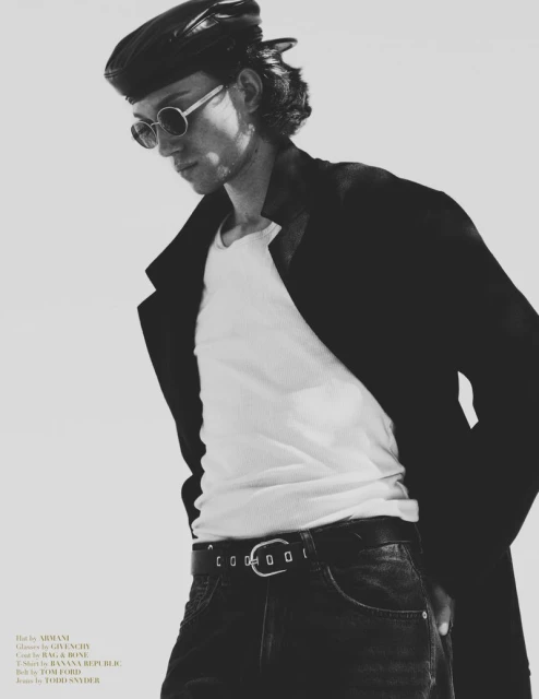 Monochrome portrait of a male fashion model in a beret, sunglasses, white ribbed t-shirt, dark jeans with a belt, and a black overcoat.