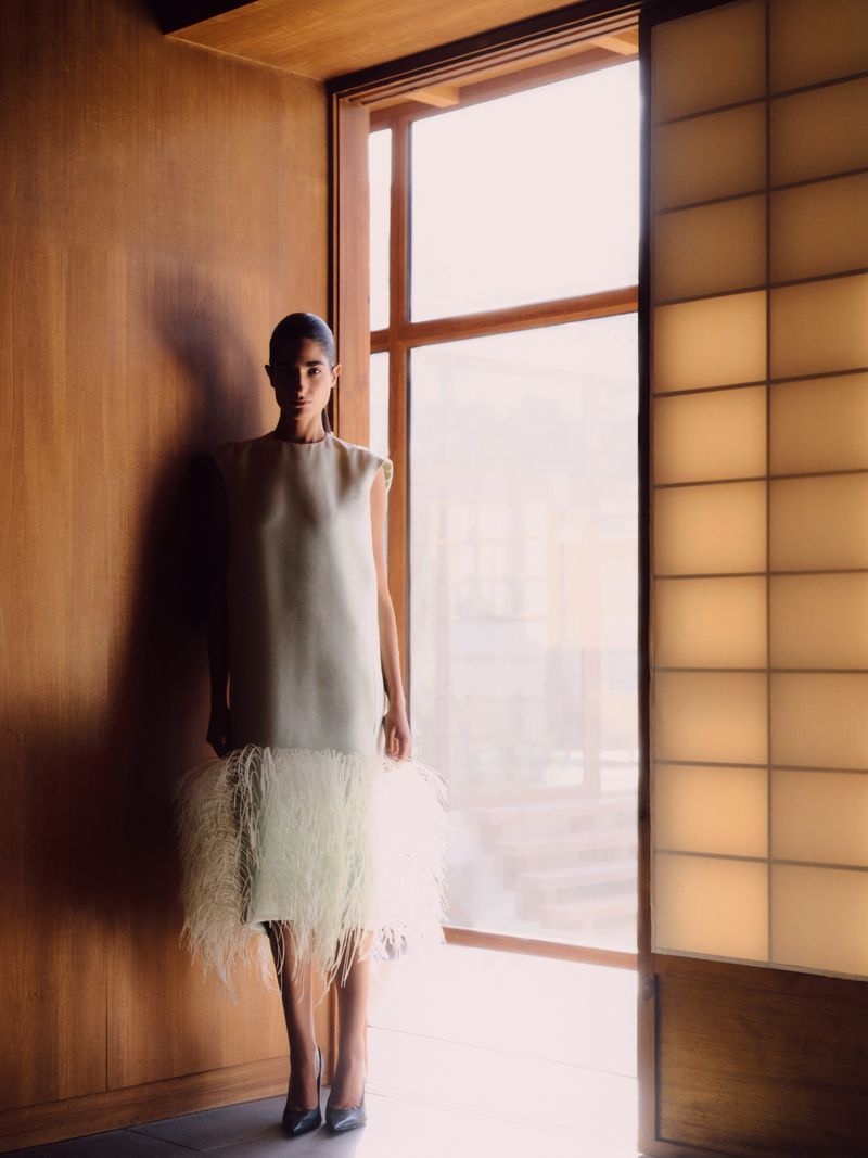 A woman in a light-colored sleeveless dress poses next to a window in a minimalist interior with wood paneled walls and a tiled doorway.
