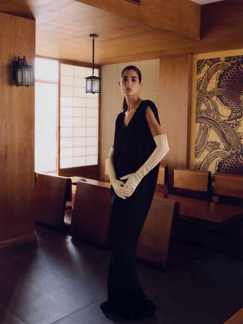 A woman poses in a black dress and long white gloves inside a paneled room with Japanese-inspired design elements and overhead lighting.
