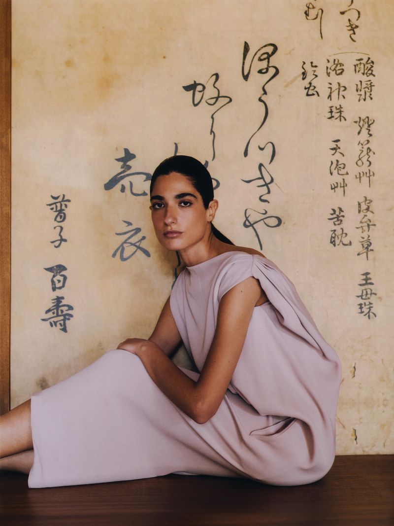 An adult female model with dark hair in a light pink dress seated in front of a wall with Japanese calligraphy in a studio setting.