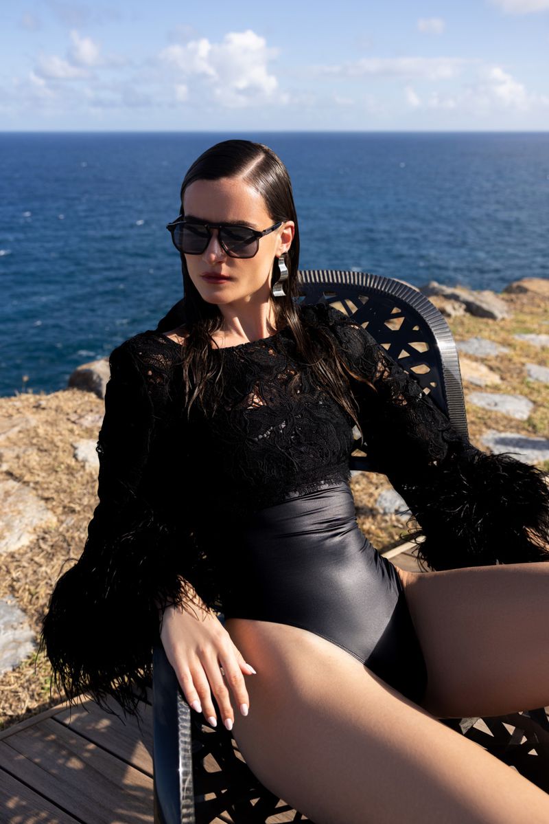 A woman wearing a black lace top and sunglasses sits in a chair outdoors with the ocean and a blue sky in the background.