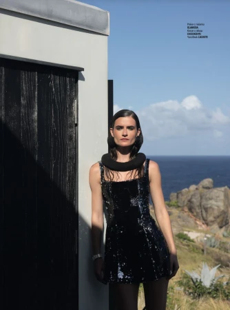 A model in a black sequined dress poses outside a white building against an ocean backdrop, under a clear blue sky in natural light.