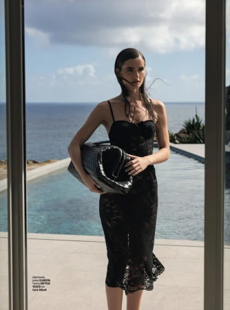Fashion shot of a woman in a black lace dress holding a large black woven handbag by an infinity pool with an ocean view.