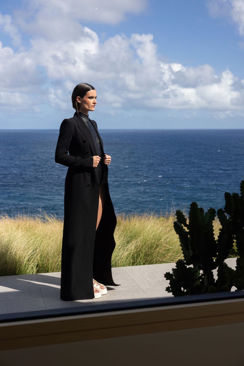 Adult female model in a black coat poses outdoors with the ocean and cloudy sky behind her, seen from a windowed balcony.