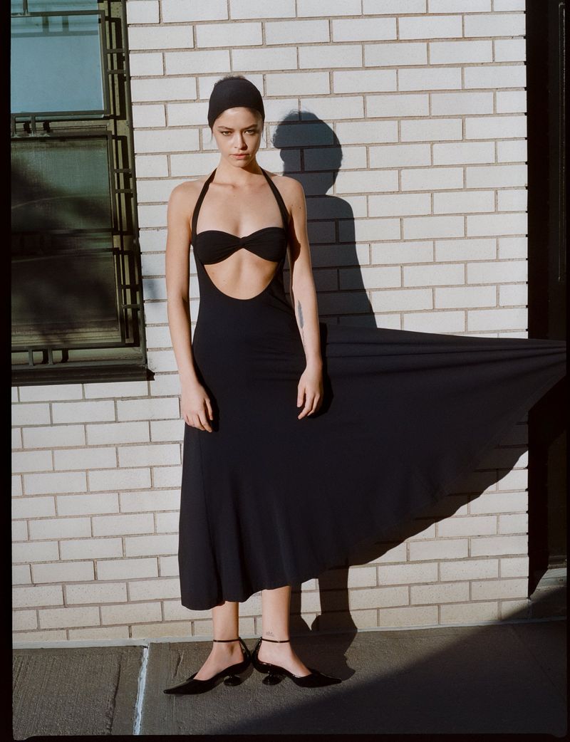 Elegant woman in a black dress and head covering poses against a brick wall in an outdoor fashion shot, ankle strap heels.