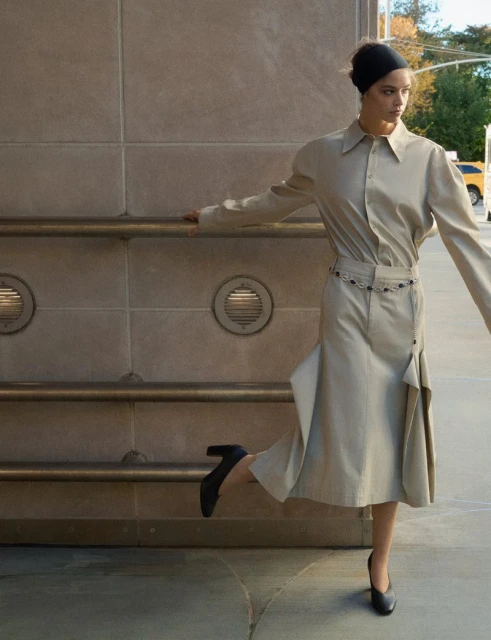 A stylish adult female with a black headband is wearing a beige shirt and skirt, standing on a city sidewalk in front of a stone wall.