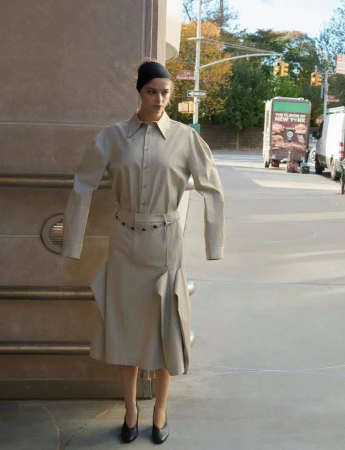 Woman in beige collared dress and black headscarf poses on urban sidewalk next to concrete building, with street and trees background.