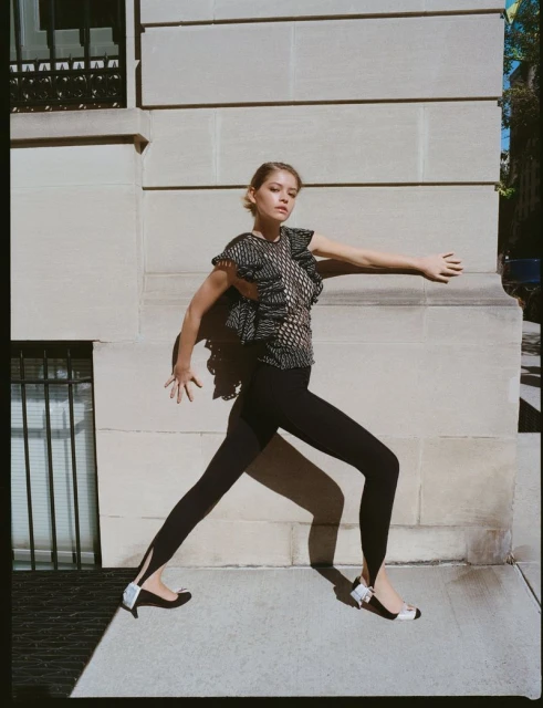 Fashion shot of a woman in a black and white ruffled top, black leggings, and ballet flats, posed in front of a stone building.