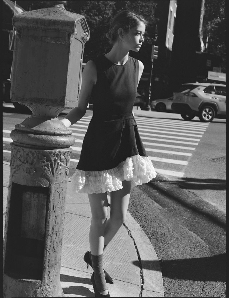 Black-and-white shot of a slender woman in a black dress with white ruffles, posing next to a decorative column in a city crosswalk.
