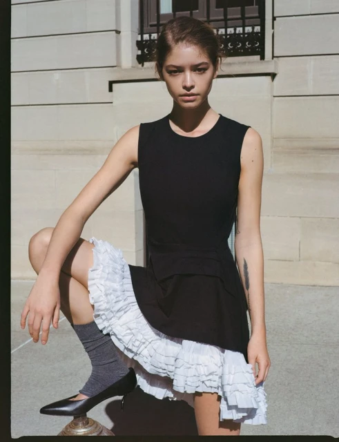 Young woman in black dress with white ruffles posing outdoors. She is sitting on a metallic surface, looking at the camera.