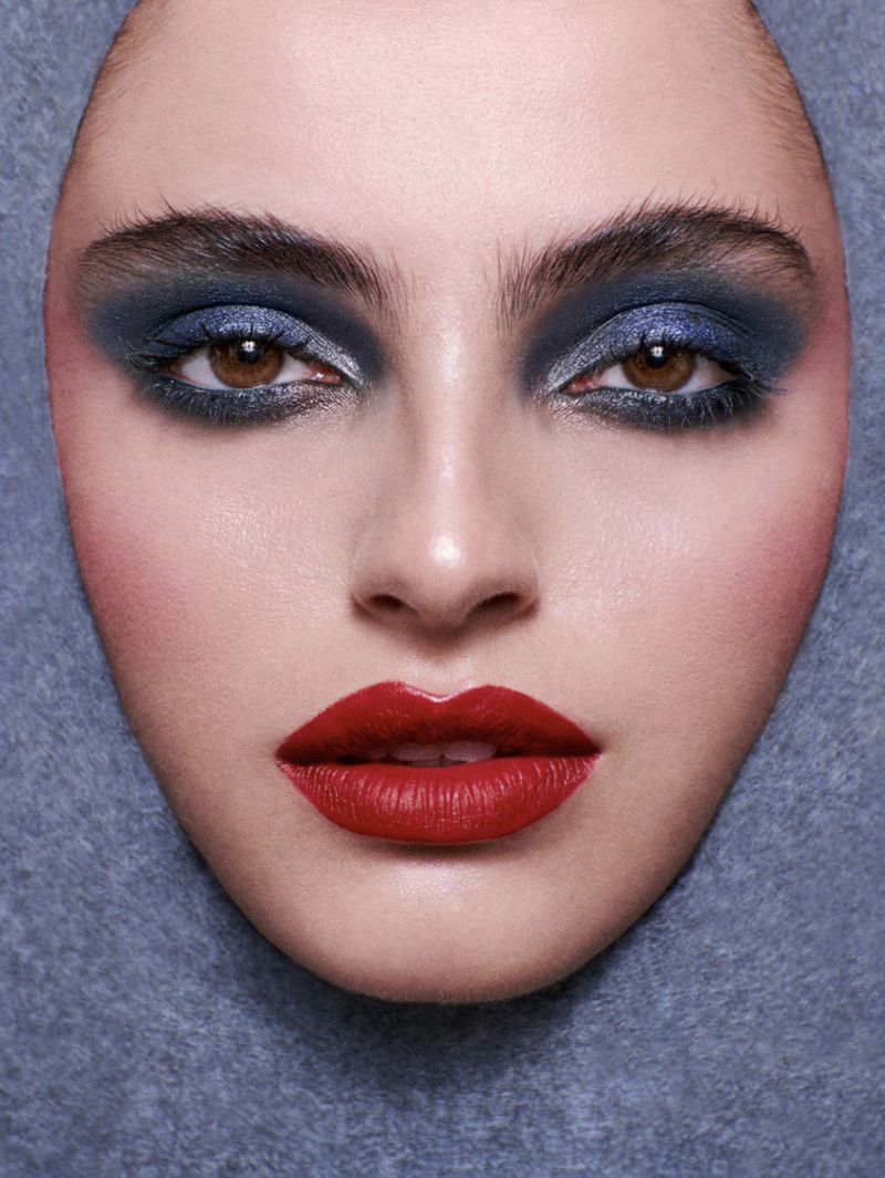 Close-up portrait of a woman's face with bold makeup featuring red lips, dark eyeshadow, and pink blush against a grey background.
