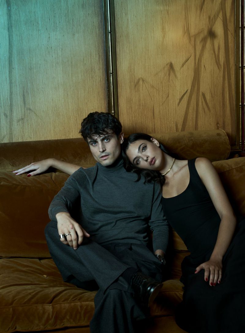 A stylish indoor portrait features a dark-haired man and woman on a couch, with a soft, diffused lighting and an editorial aesthetic.