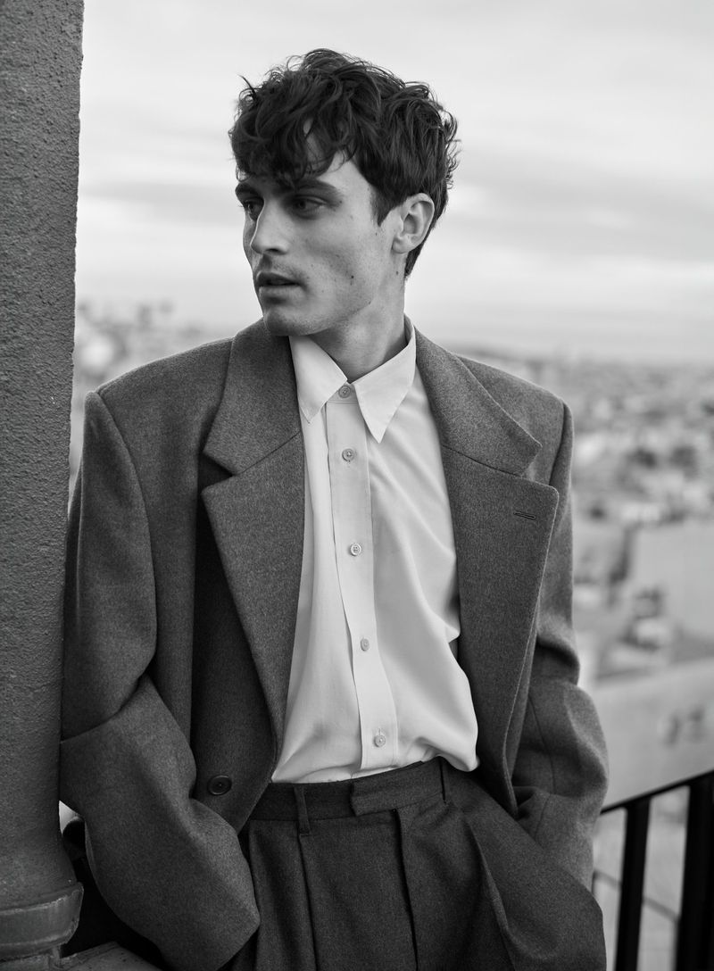 Monochrome portrait of a young adult man in overcoat and button-down shirt, hands in pockets, gazing away in an urban environment.