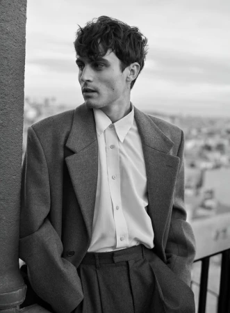 Monochrome portrait of a young adult man in overcoat and button-down shirt, hands in pockets, gazing away in an urban environment.