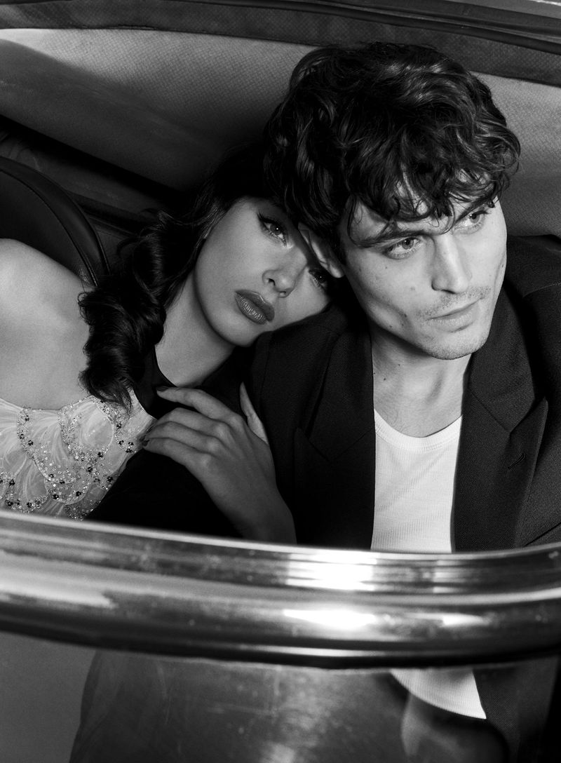 Black-and-white shot of a fashionable couple indoors, with the woman leaning on the man's shoulder, both looking elegant and stylish.