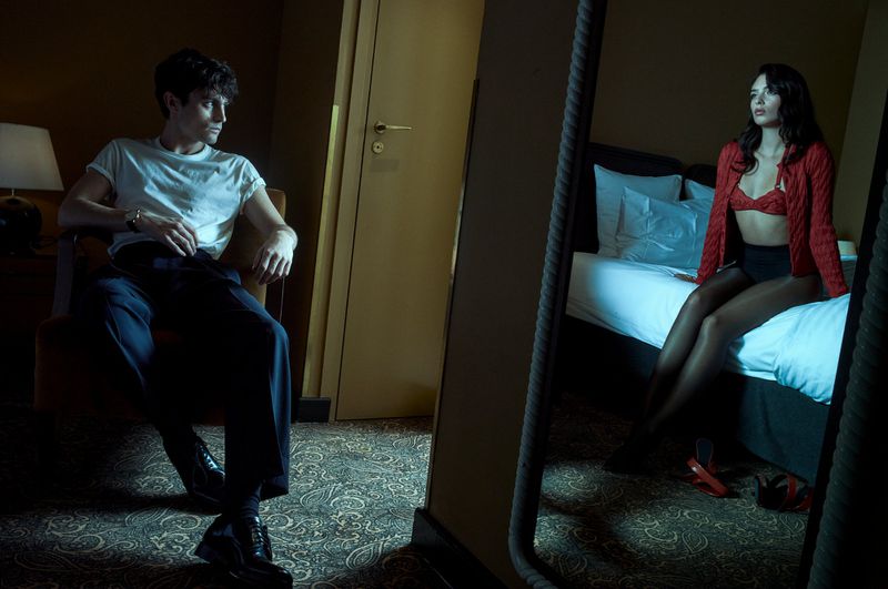 Man and woman in a bedroom setting with a mirror reflection showing the woman in lingerie, red cardigan, and black hosiery.
