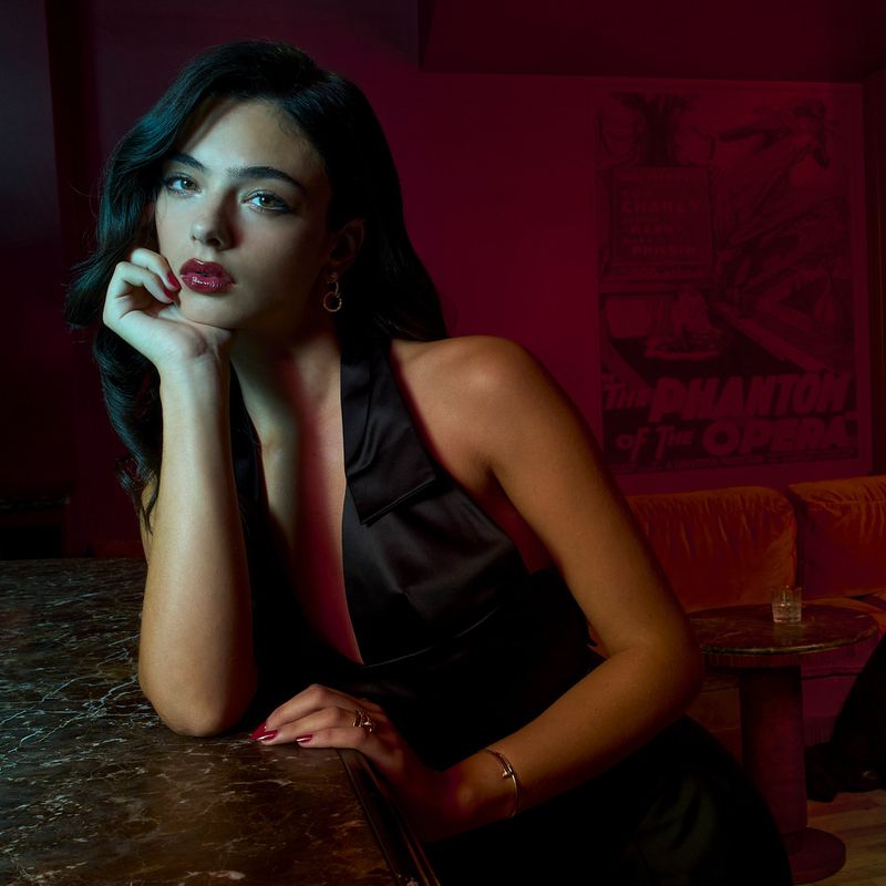 Portrait of a young woman with dark hair and red lipstick leaning on a marble countertop, wearing a black dress in moody lighting.