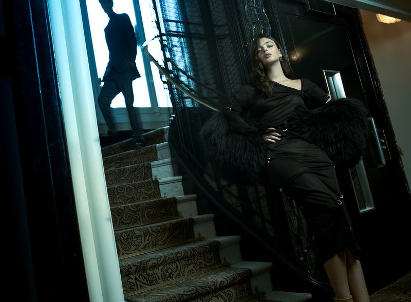 Elegant woman in black dress and fur stole poses on staircase with patterned carpet, while a blurred man ascends in the background.