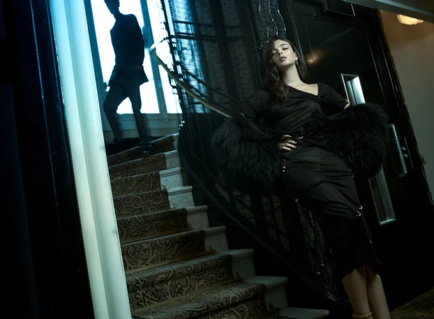 Elegant woman in black dress and fur stole poses on staircase with patterned carpet, while a blurred man ascends in the background.