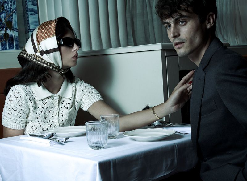 Woman in headscarf and sunglasses sits at a table with a man in a suit; they're in a restaurant with dishes and drinks present.