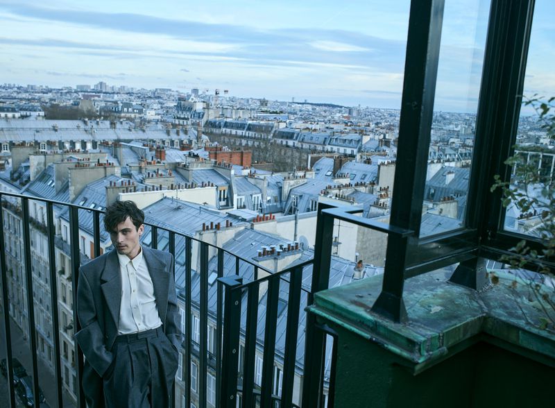 Young man in grey suit poses on a balcony with a cityscape of Paris rooftops in the background under a cloudy sky.