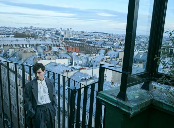 Young man in grey suit poses on a balcony with a cityscape of Paris rooftops in the background under a cloudy sky.