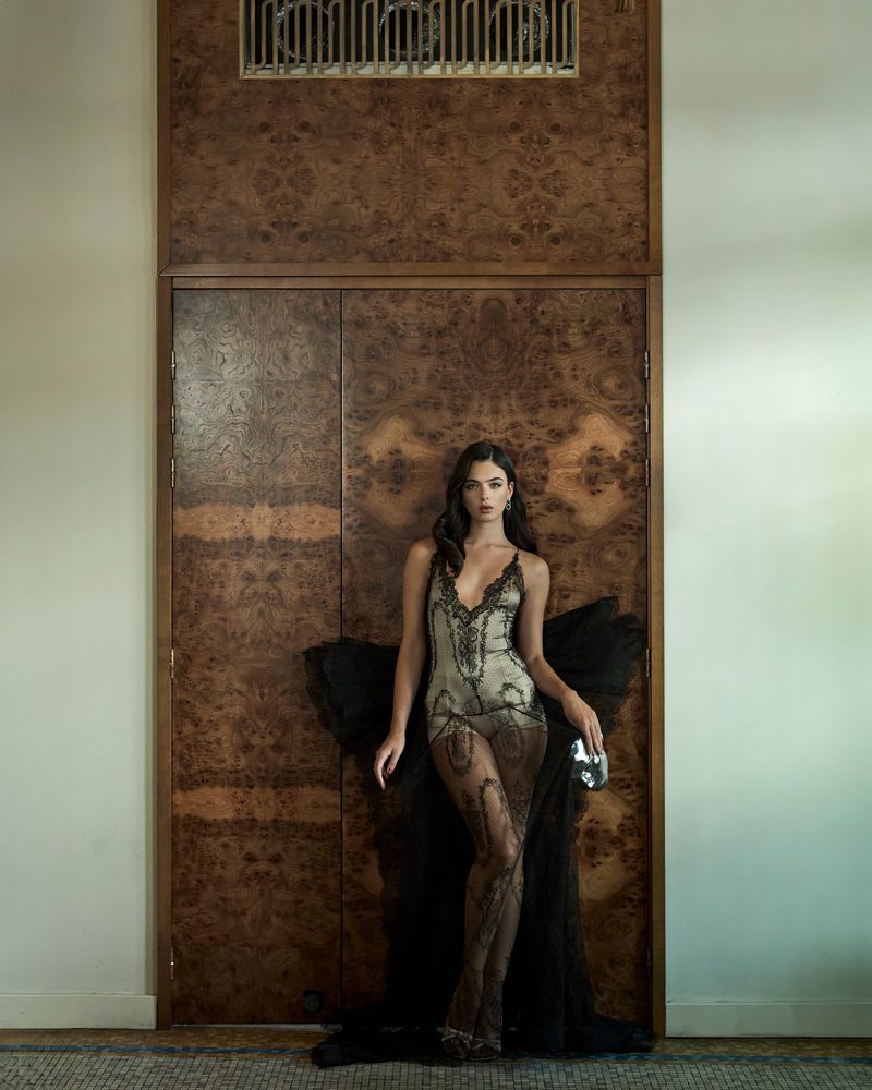 Elegant woman in a beaded dress with a black tulle train poses against a textured wooden wall in an indoor fashion setting.