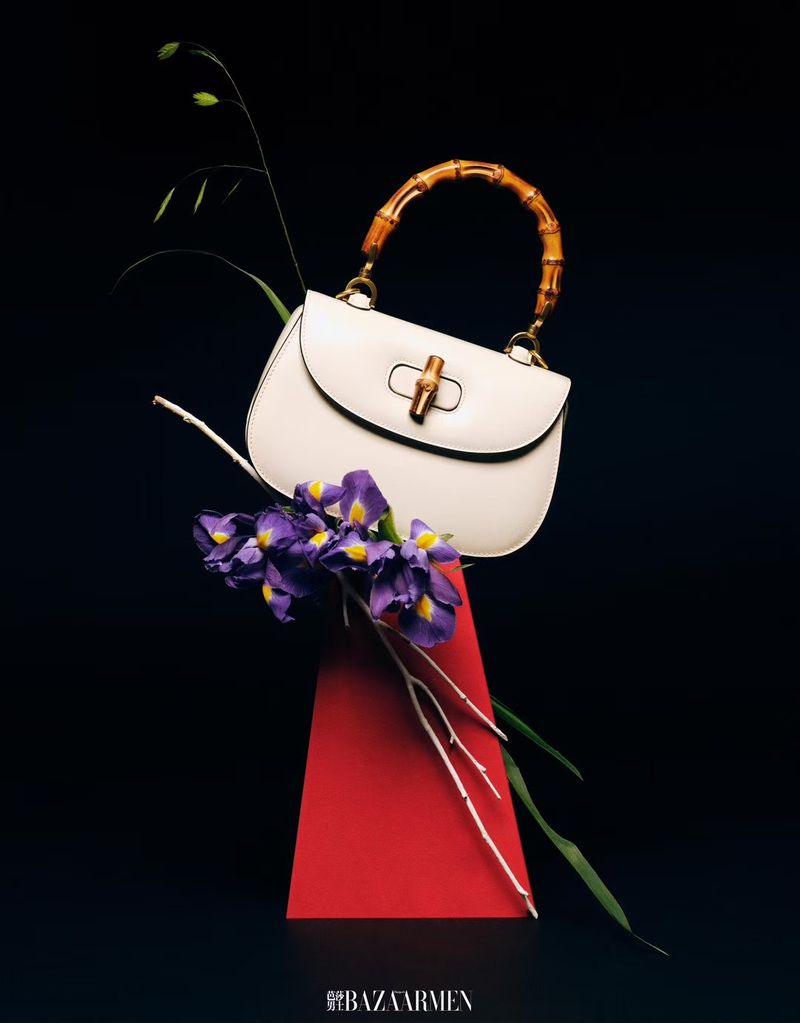 Fashion shot of a white leather purse with bamboo handle, purple flowers, and a red geometric shape against a black background.