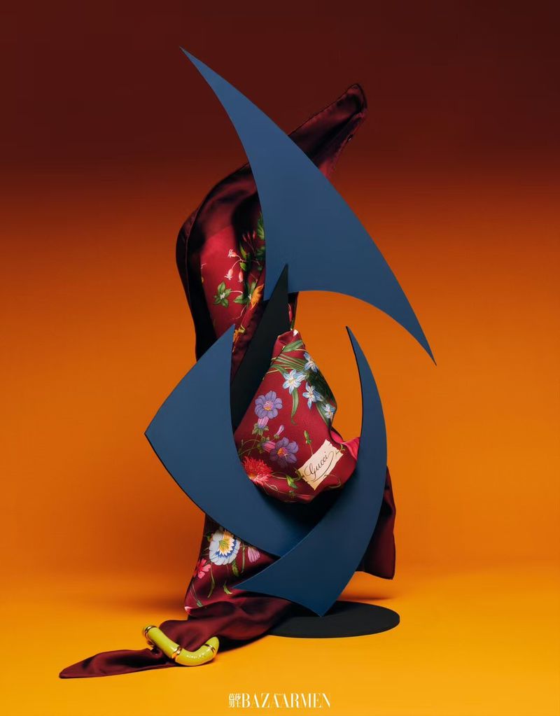 Still life featuring a silk scarf with floral design and a blue geometric sculpture against a gradient orange background.