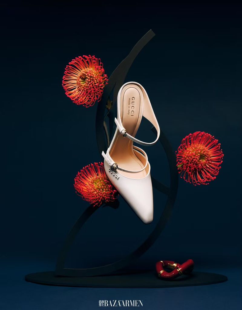 Cream-colored Gucci heeled sandal with flowers and sculptural stand, photographed in a still life composition against a dark blue background.