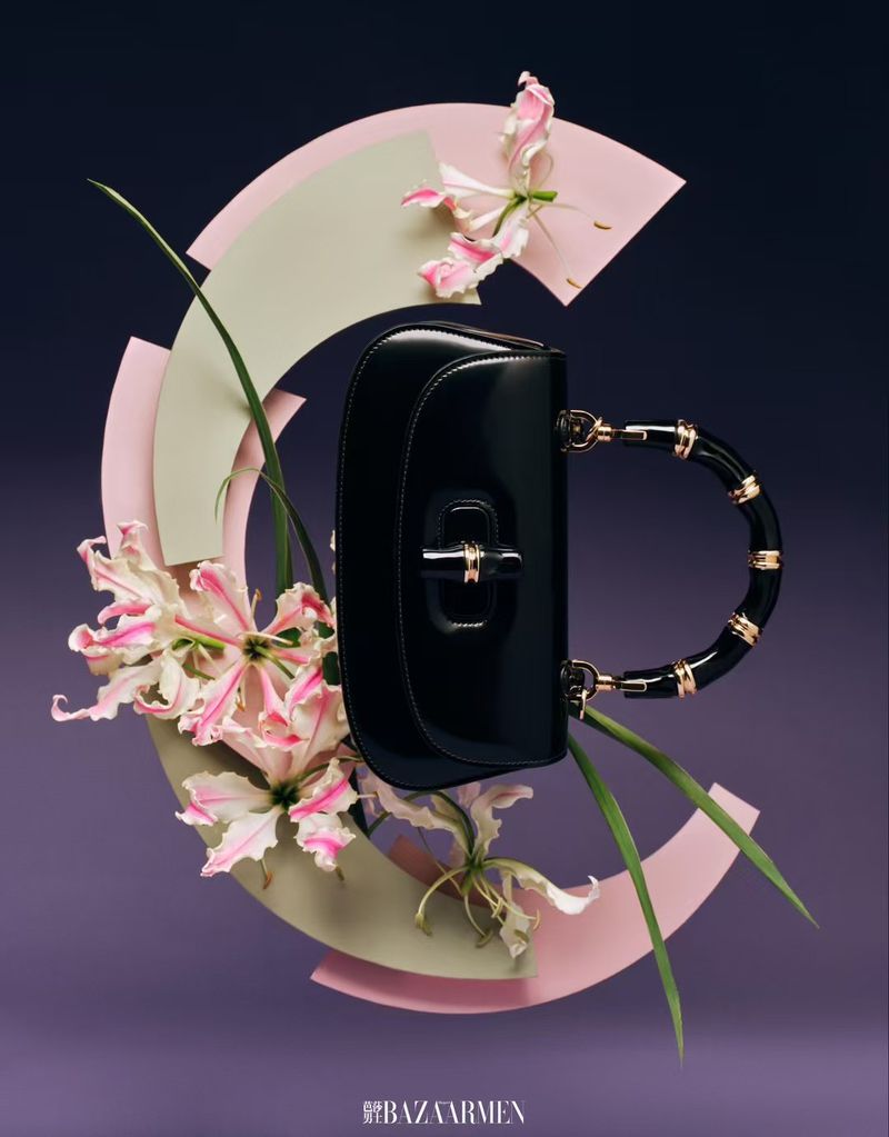 Elegant black handbag with a distinctive handle design, set against a backdrop of pastel geometric shapes and floral accents in a studio setting.
