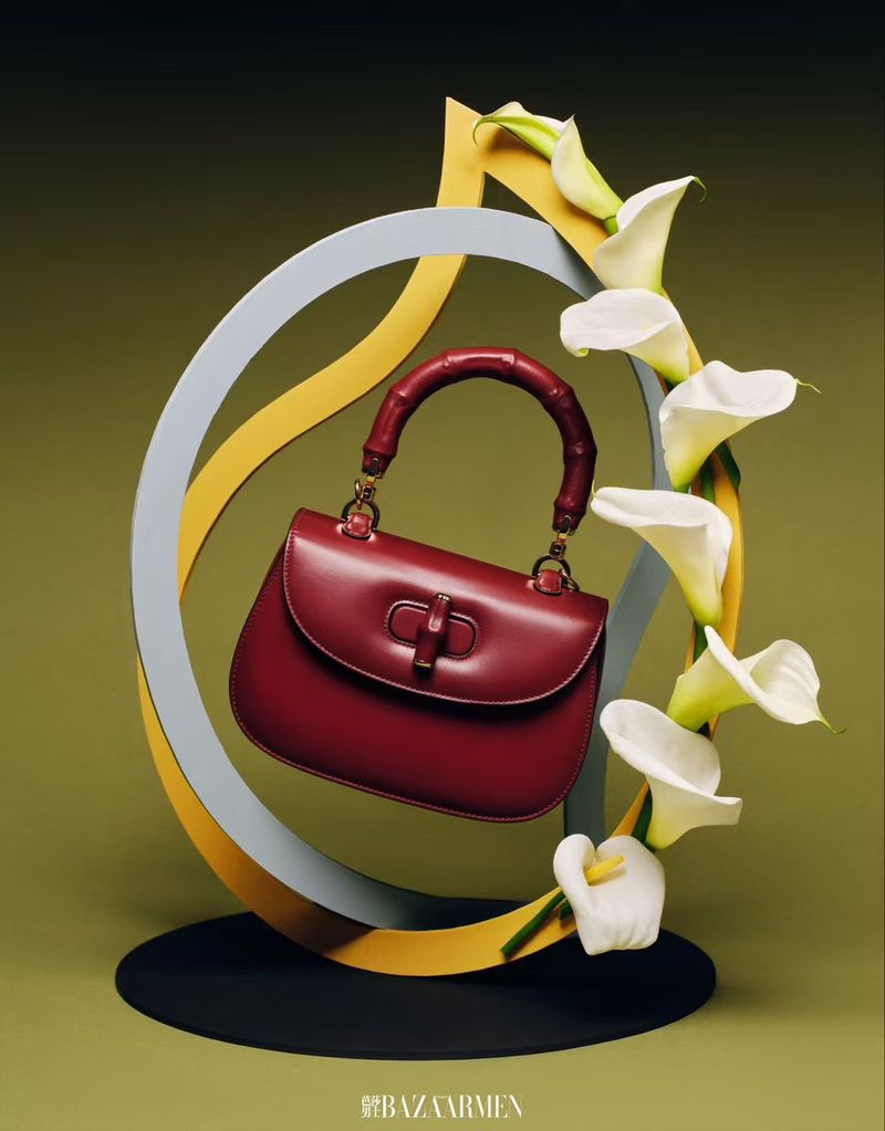 Burgundy leather handbag with bamboo handle is displayed with white calla lilies against a muted green backdrop.