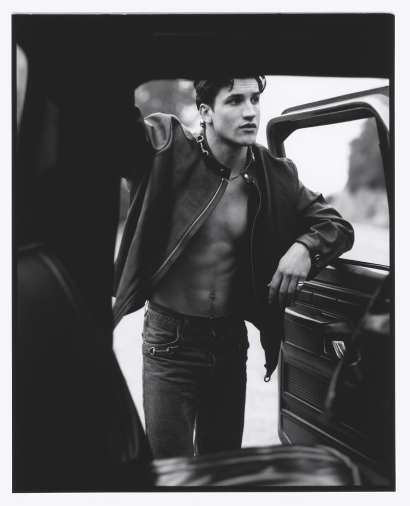 Monochrome portrait of a young adult male with an open jacket, standing by a vehicle in an outdoor setting with natural light.