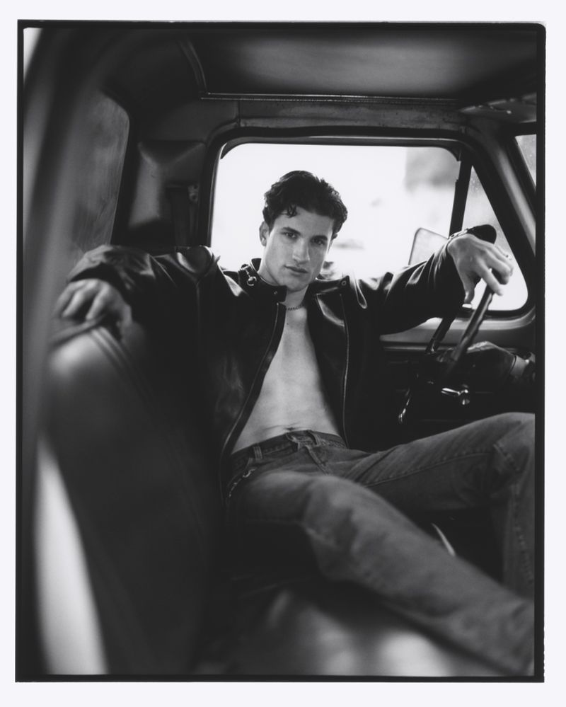 Monochrome portrait of a muscular adult male sitting in a car, wearing a leather jacket and jeans, showcasing a relaxed pose and intense gaze.