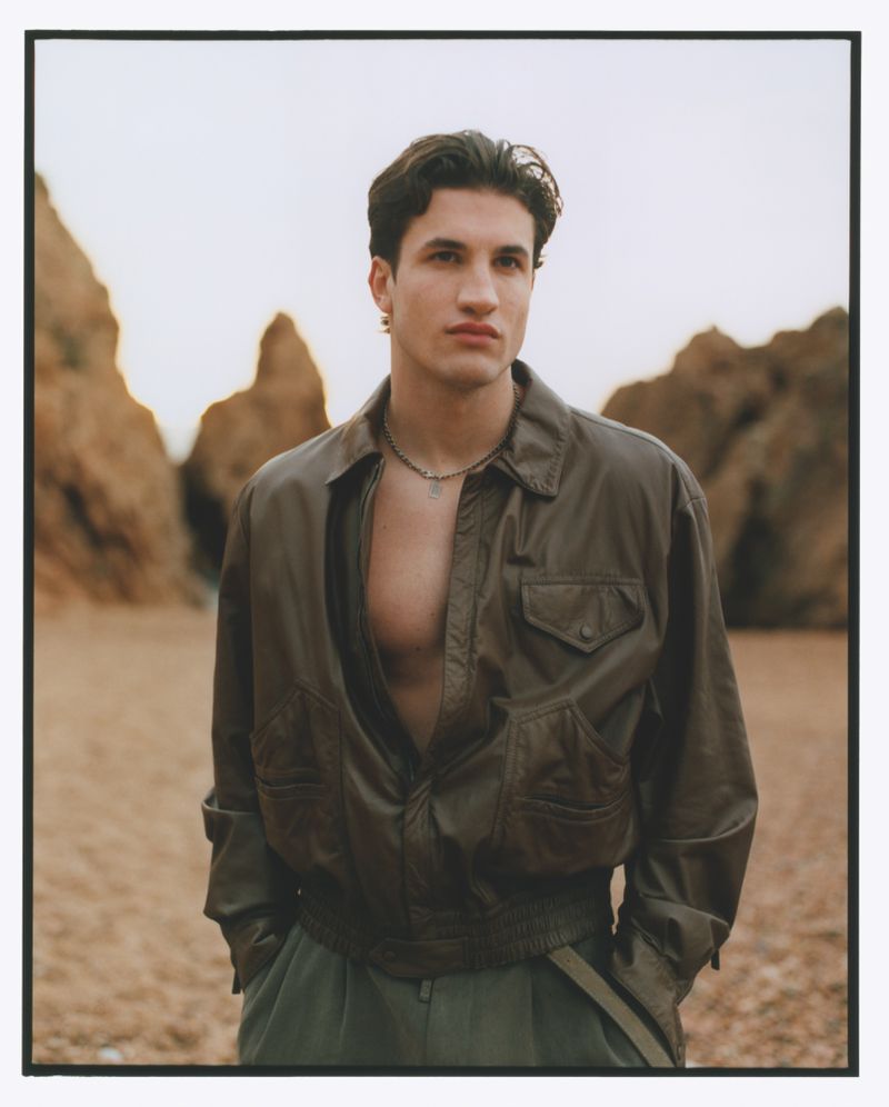 Portrait of a dark-haired man in an unbuttoned olive jacket and green pants standing on a beach with cliffs in the background.