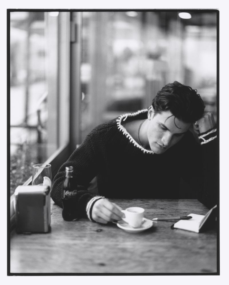 Young man in a sweater sitting at a table in a cafe, drinking coffee and writing in a notebook, with a Coca-Cola bottle nearby.