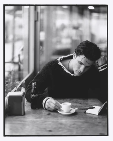 Young man in a sweater sitting at a table in a cafe, drinking coffee and writing in a notebook, with a Coca-Cola bottle nearby.