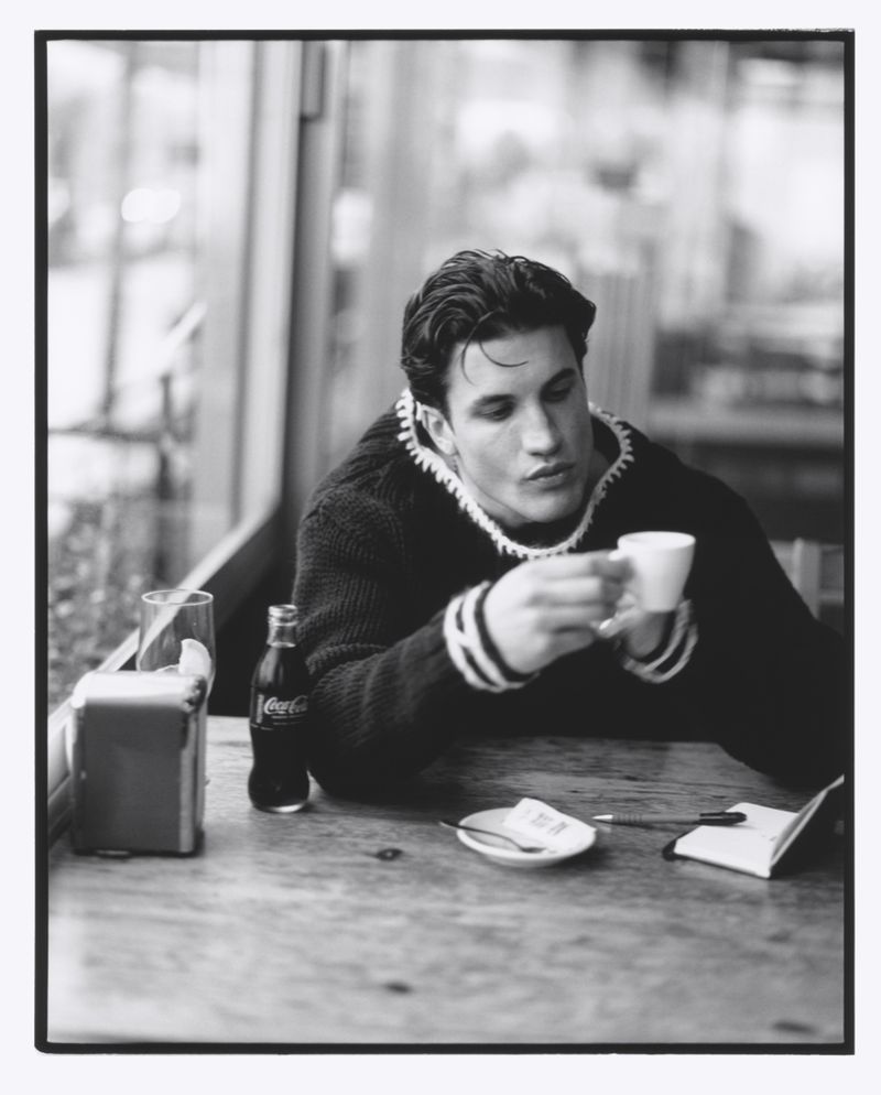 Black and white portrait of a man with dark hair sitting at a cafe table, holding a coffee cup, with a Coca-Cola bottle nearby.