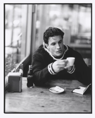 Black and white portrait of a man with dark hair sitting at a cafe table, holding a coffee cup, with a Coca-Cola bottle nearby.