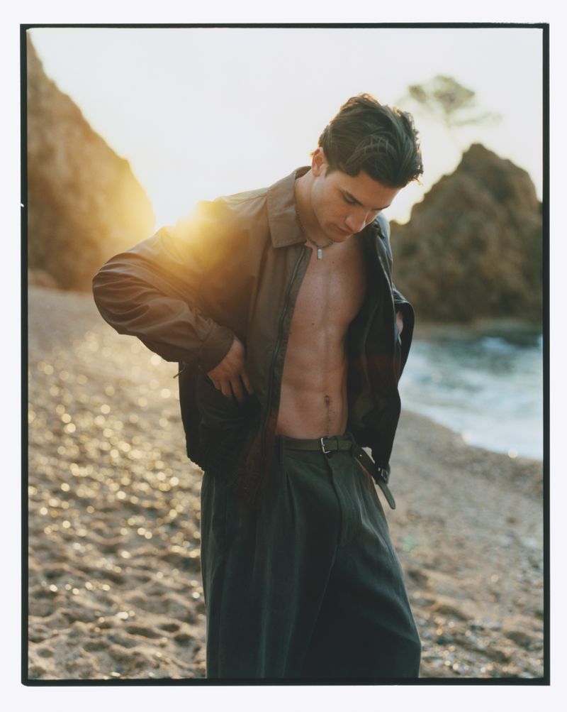 A young man on a beach at sunset, wearing an olive jacket, no shirt, and olive slacks. He is looking down with mountains in the background.