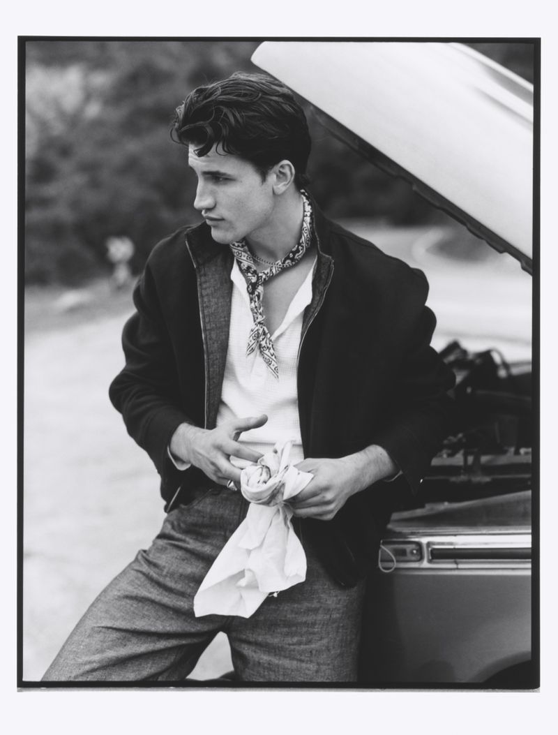 Monochrome portrait of a young adult male wearing a jacket and neck scarf, leaning against a car, with greenery in the background.