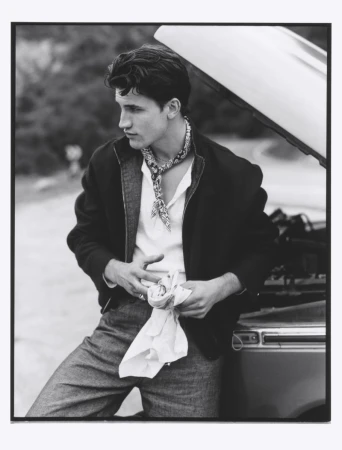 Monochrome portrait of a young adult male wearing a jacket and neck scarf, leaning against a car, with greenery in the background.