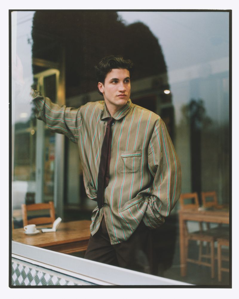 Portrait of a young adult male with dark hair, wearing a striped shirt and tie, leaning against a reflective window, looking away.